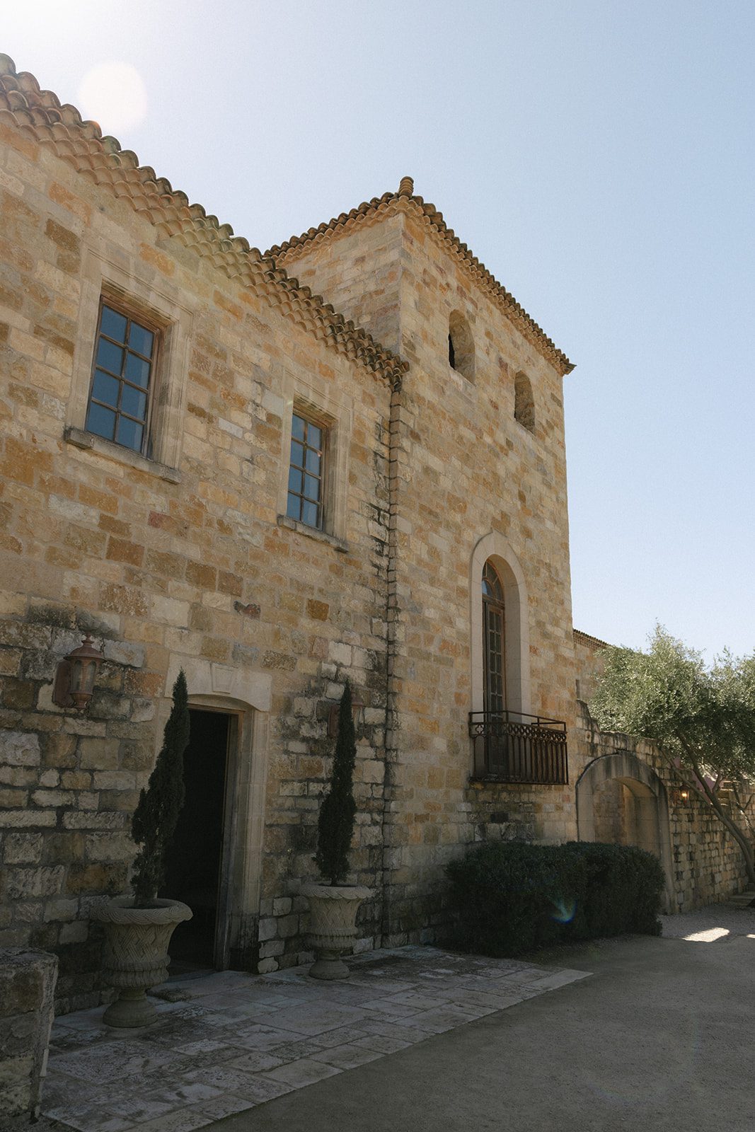 Sunlit exterior of Sunstone Villa in Santa Ynez, California, showcasing the wedding venue's European-inspired limestone architecture, terracotta roof, arched doorway, and elegant courtyard surrounded by olive trees.