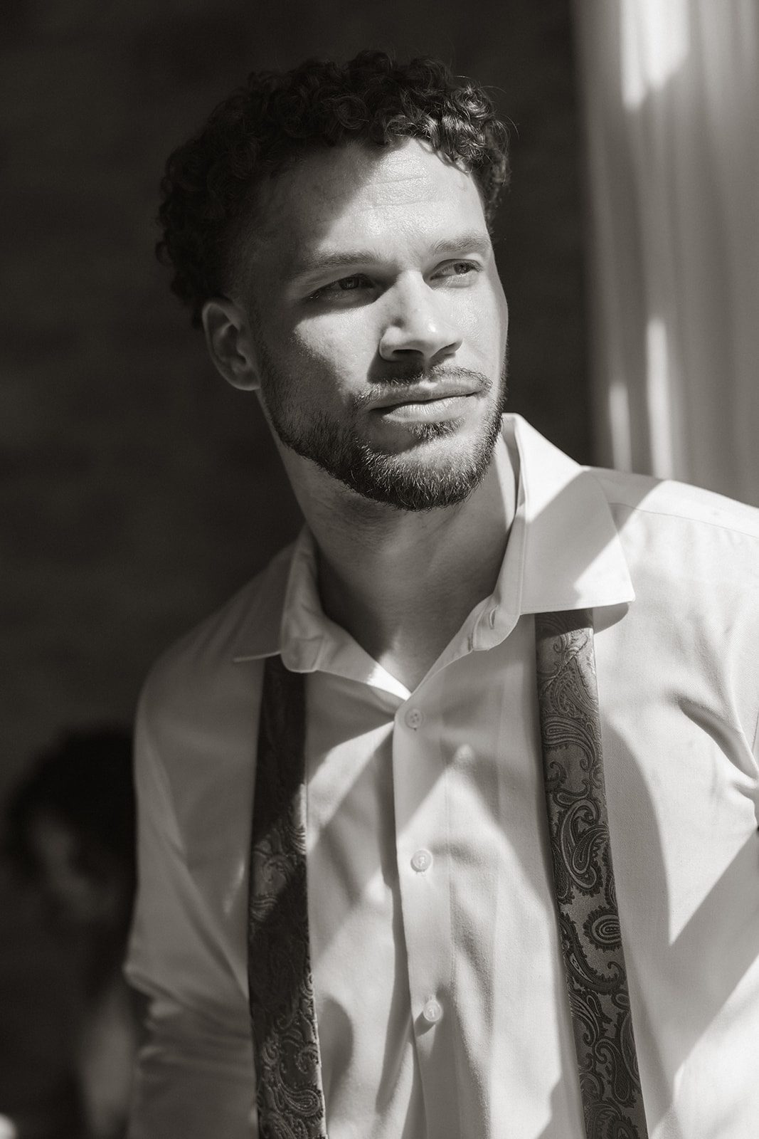 Black and white editorial portrait of a groom wearing an unbuttoned white dress shirt and patterned tie, captured in dramatic window light by a California wedding photographer.