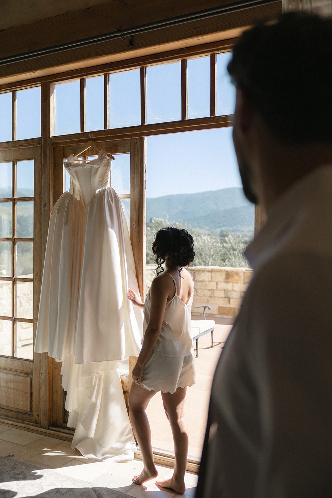 Bride admiring her strapless satin wedding gown hanging beside rustic wooden French doors, with Santa Ynez Valley hills visible beyond the stone terrace at Sunstone Villa.