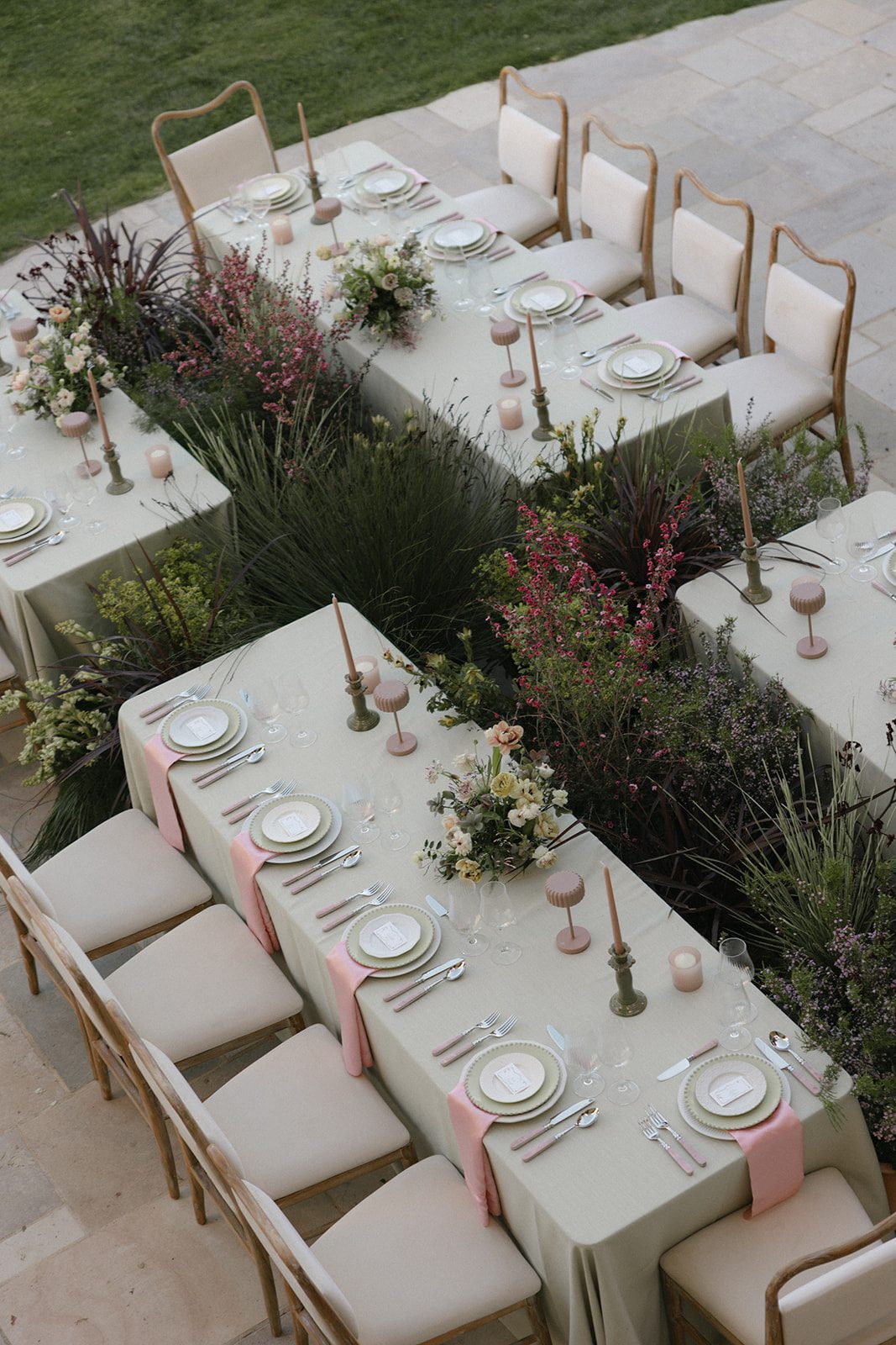 Overhead view of an elegant outdoor wedding reception at Sunstone Villa, featuring sage green table linens, blush pink napkins, garden-inspired floral arrangements, vintage wooden chairs, and lush greenery between the tables.
