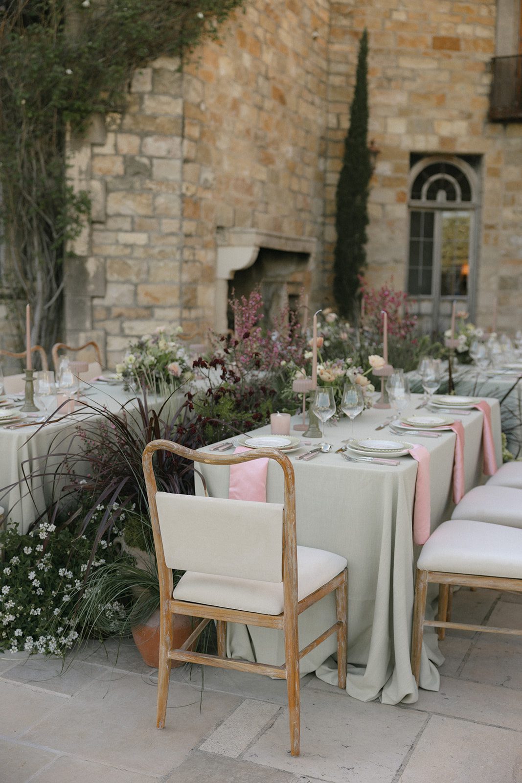 Romantic outdoor wedding reception at Sunstone Villa in Santa Ynez, featuring sage green table linens, blush pink napkins, garden-style floral centerpieces, and wooden chairs against a rustic stone backdrop.