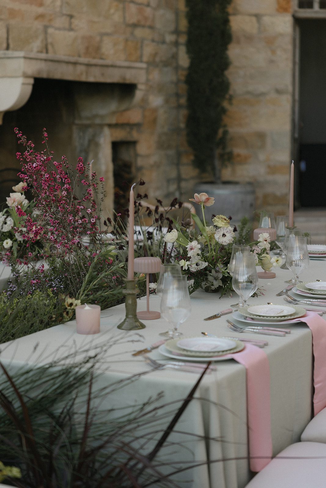Romantic wedding reception tablescape at Sunstone Villa in Santa Ynez, featuring delicate pink and ivory flowers, sage green linens, blush pink napkins, vintage candlesticks, and layered place settings against rustic stone architecture.