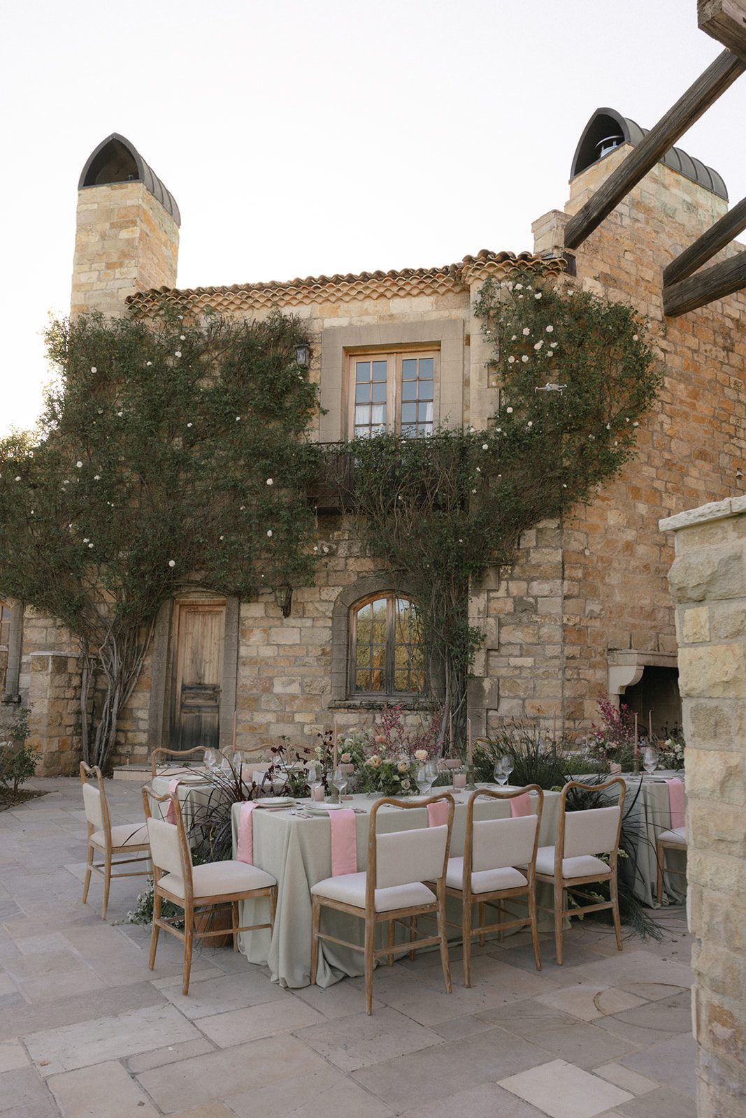 European inspired wedding reception at Sunstone Villa featuring long dining tables, soft pink linens, garden-style floral arrangements, and rustic wooden chairs beneath the villa's stone facade.