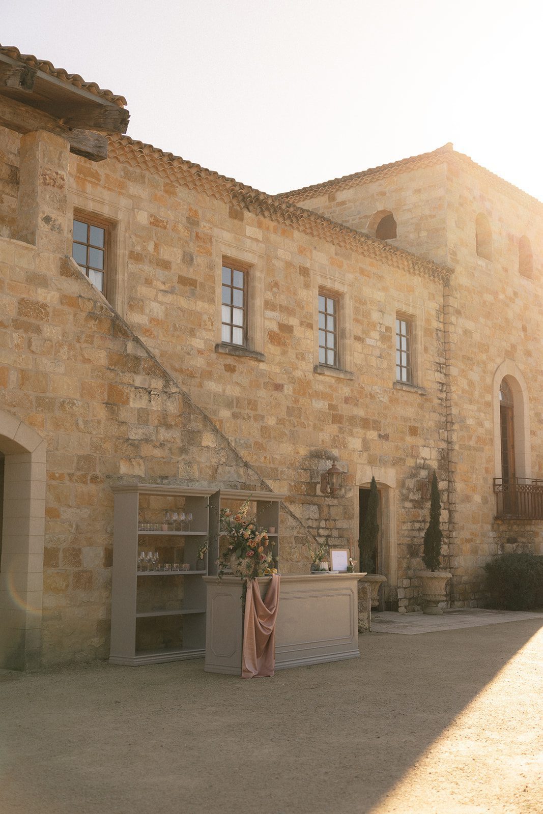 Golden afternoon sunlight illuminating the French-inspired stone architecture of Sunstone Villa, with an elegant wedding bar decorated in dusty rose fabric and lush floral arrangements.
