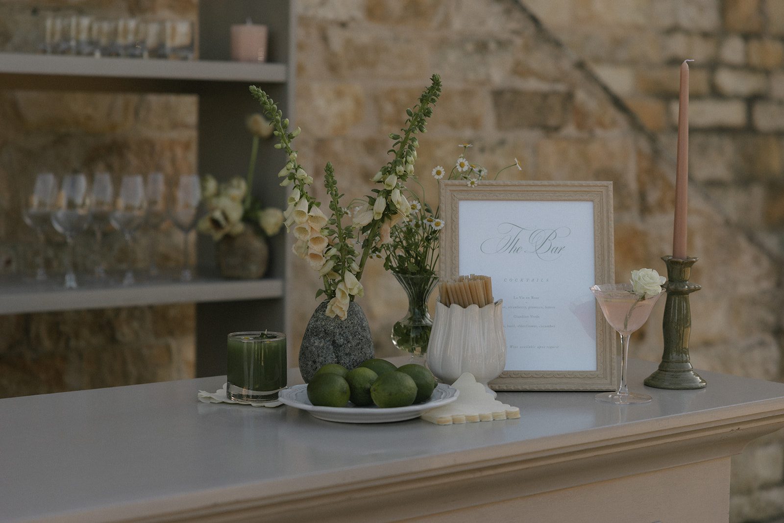 Romantic wedding cocktail bar at Sunstone Villa in Santa Ynez, featuring a framed signature cocktail menu, pink martini, fresh limes, garden-style floral arrangements, vintage green candlestick, and rustic limestone backdrop.