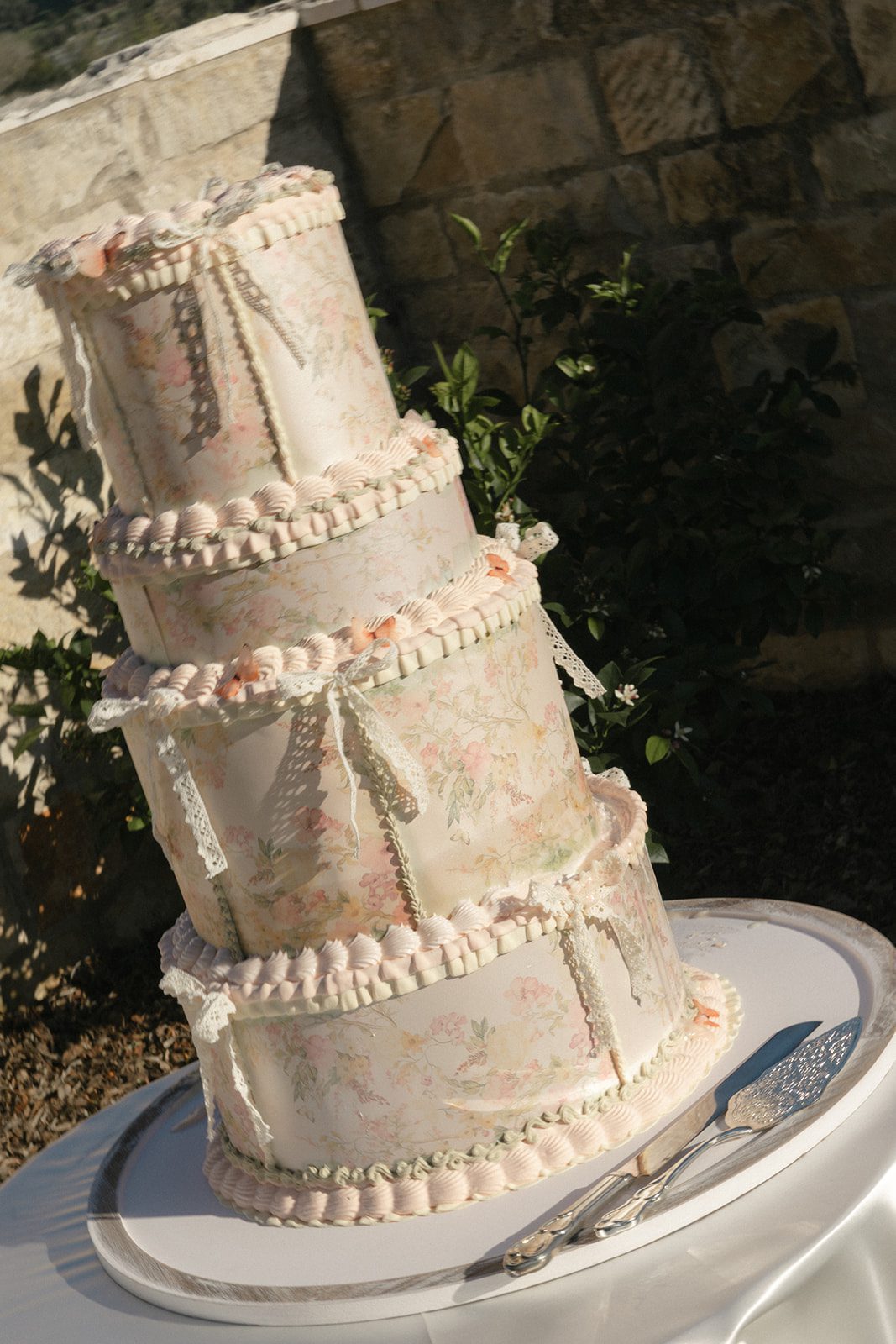 Whimsical four-tier wedding cake featuring delicate pastel floral patterns, vintage-inspired pink piping, and lace bows, photographed in warm sunlight at Sunstone Villa in California.