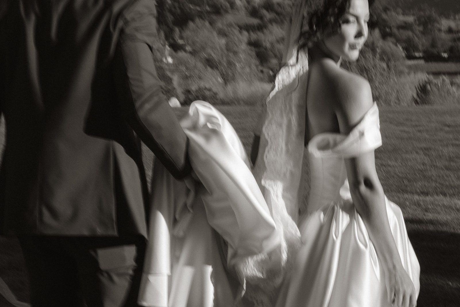Black and white candid wedding photograph of a bride in an off-the-shoulder satin gown and lace veil walking beside her groom, who carries part of her dress.