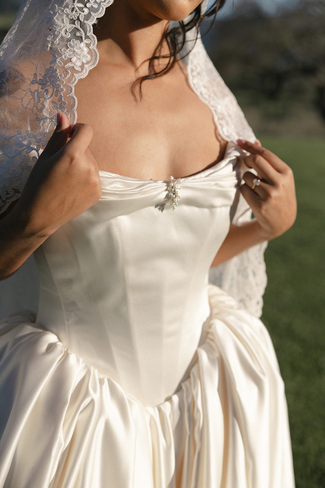 Close-up bridal fashion portrait highlighting an ivory satin wedding gown with a structured corset bodice, delicate pearl embellishment, and romantic lace mantilla veil, photographed in warm California golden hour sunlight.