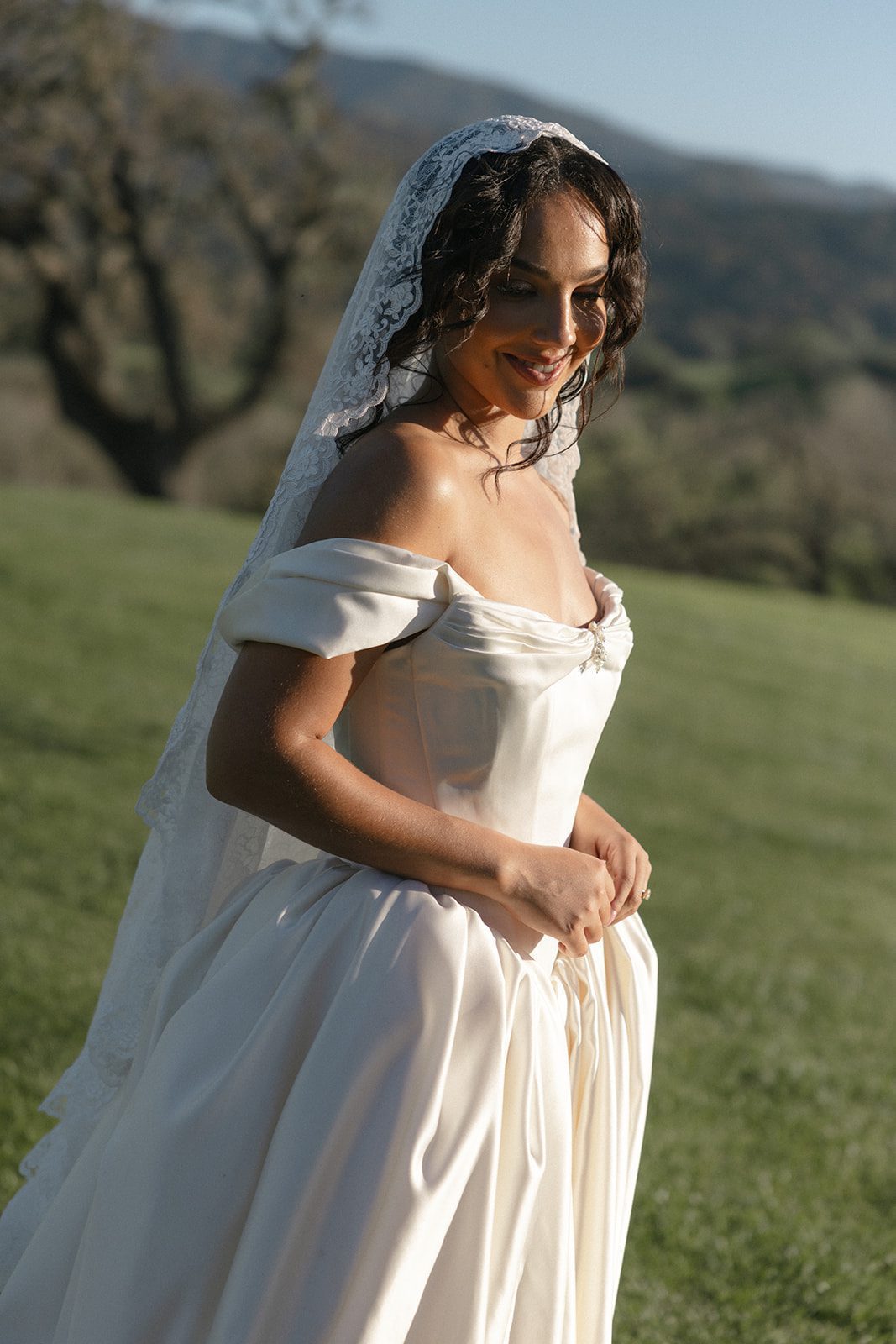 Romantic golden hour bridal portrait at Sunstone Villa in Santa Ynez, featuring a bride in an ivory off-the-shoulder satin wedding gown and delicate lace mantilla veil, with rolling California hills in the background.