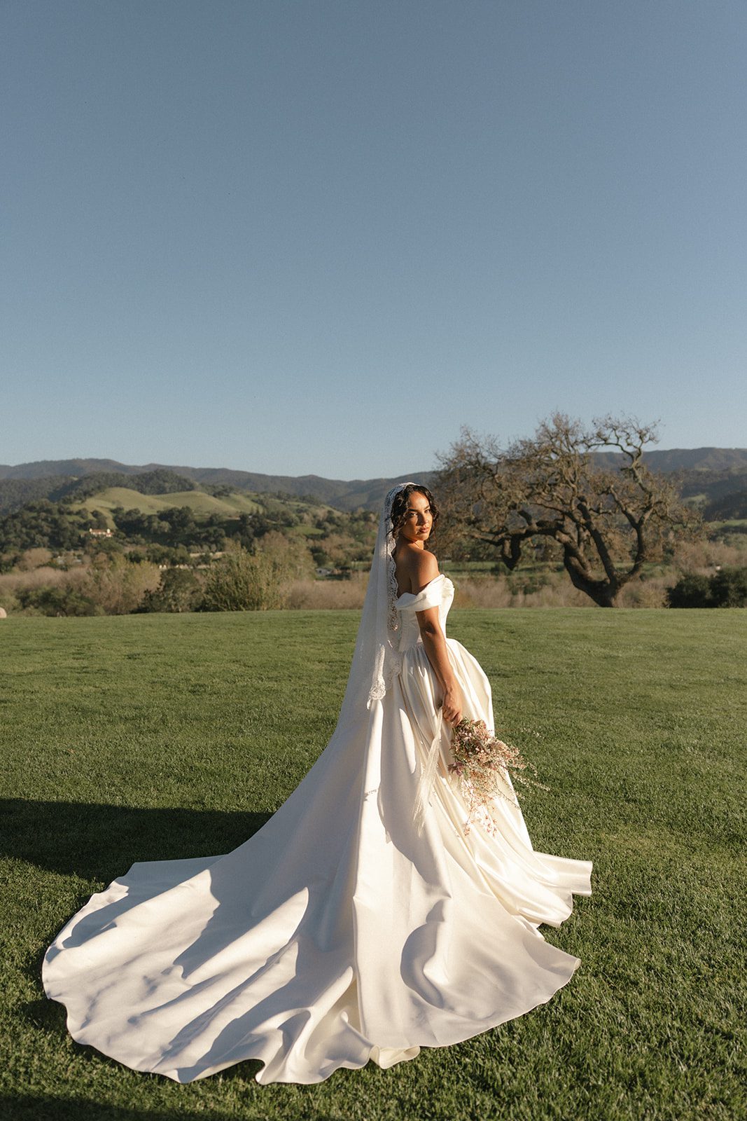 Golden hour bridal portrait at Sunstone Villa in Santa Ynez, California, featuring a bride in a flowing ivory satin wedding gown and lace mantilla veil holding a romantic garden-style bouquet against rolling green hills and oak trees.
