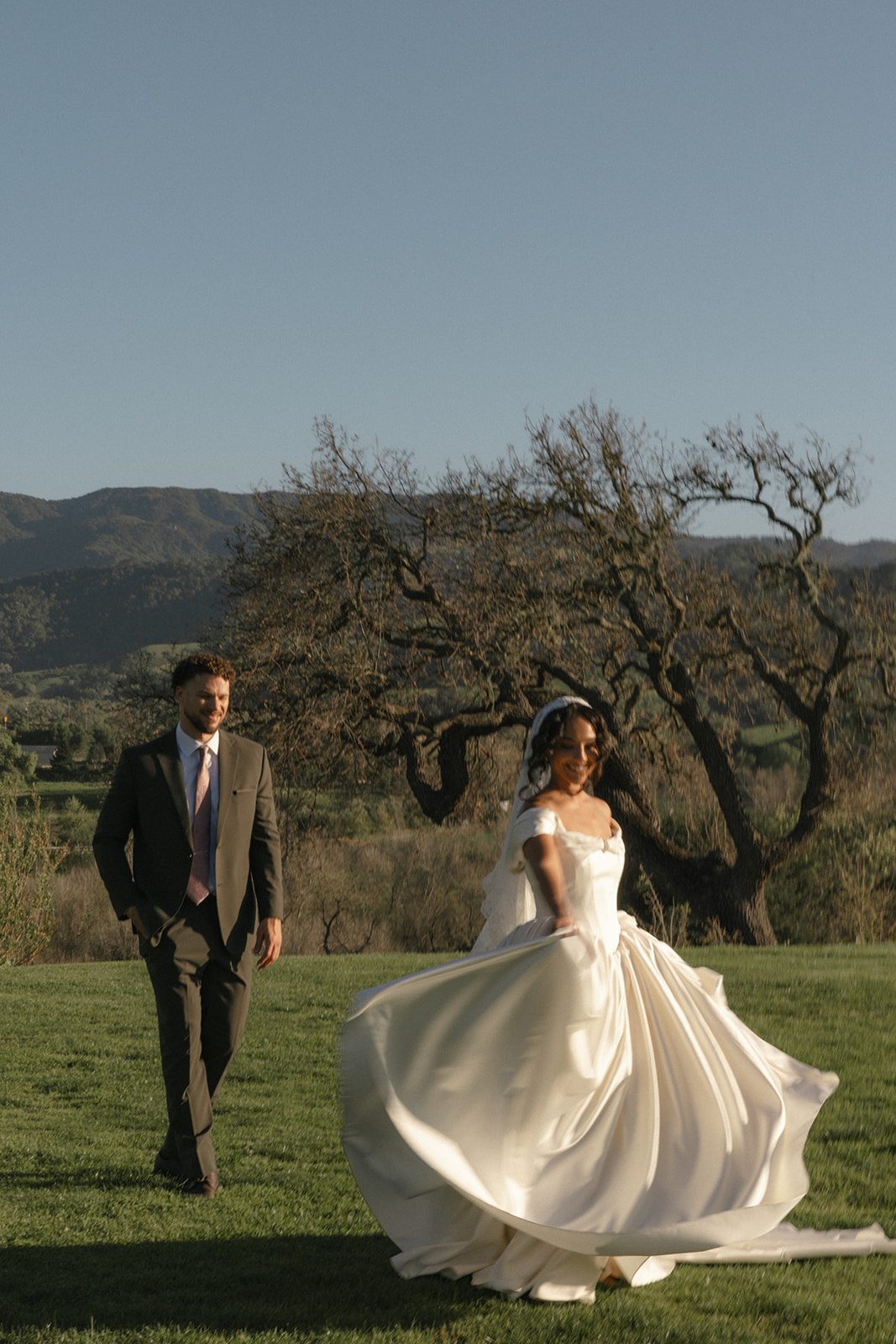 Bride twirling in her flowing satin wedding gown as the groom walks behind her during golden hour at their Sunstone Villa Wedding in Santa Ynez, California.