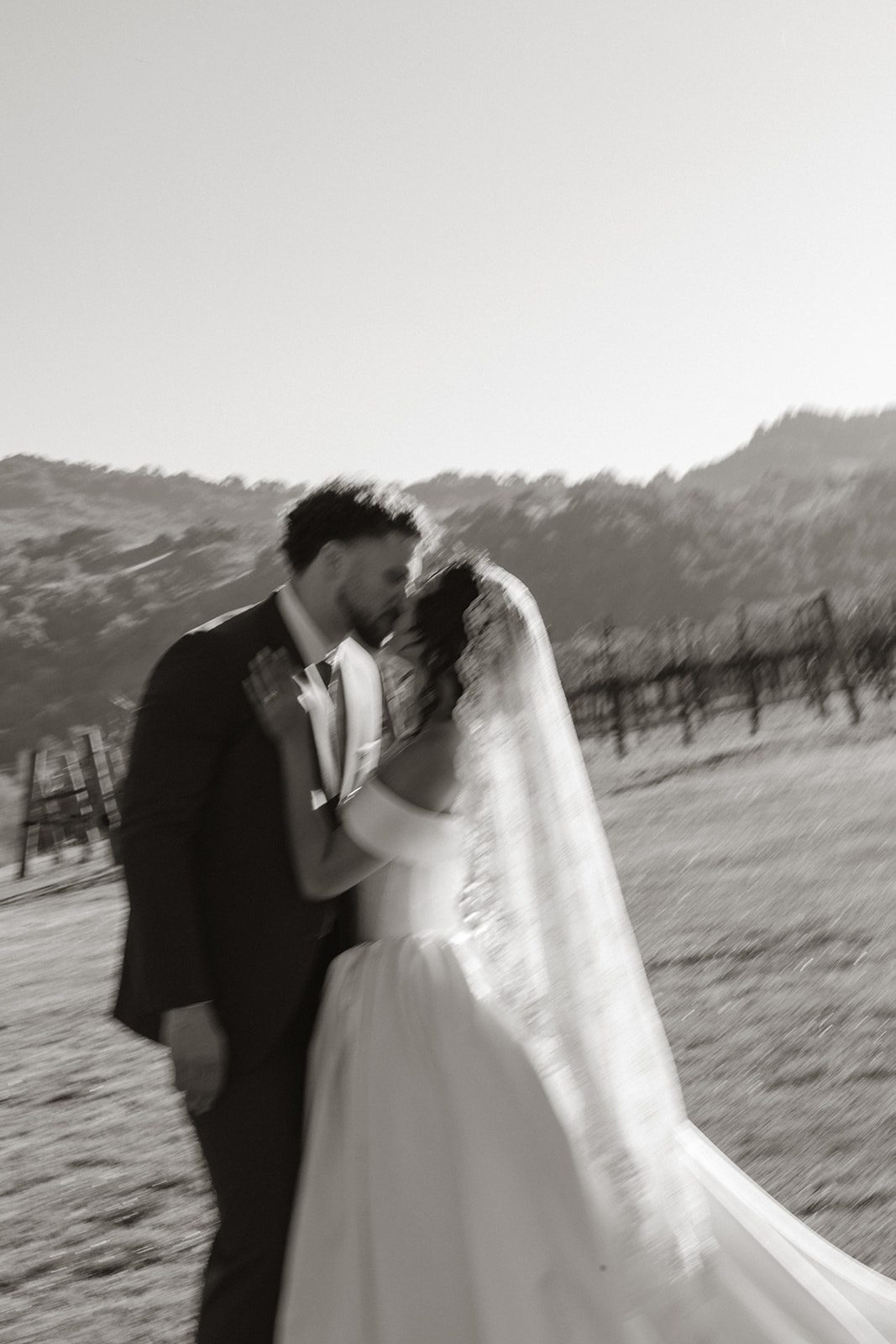 Motion-blurred black and white portrait of a bride and groom kissing beside the vines during their Sunstone Villa Wedding, with her lace veil flowing in the afternoon light.