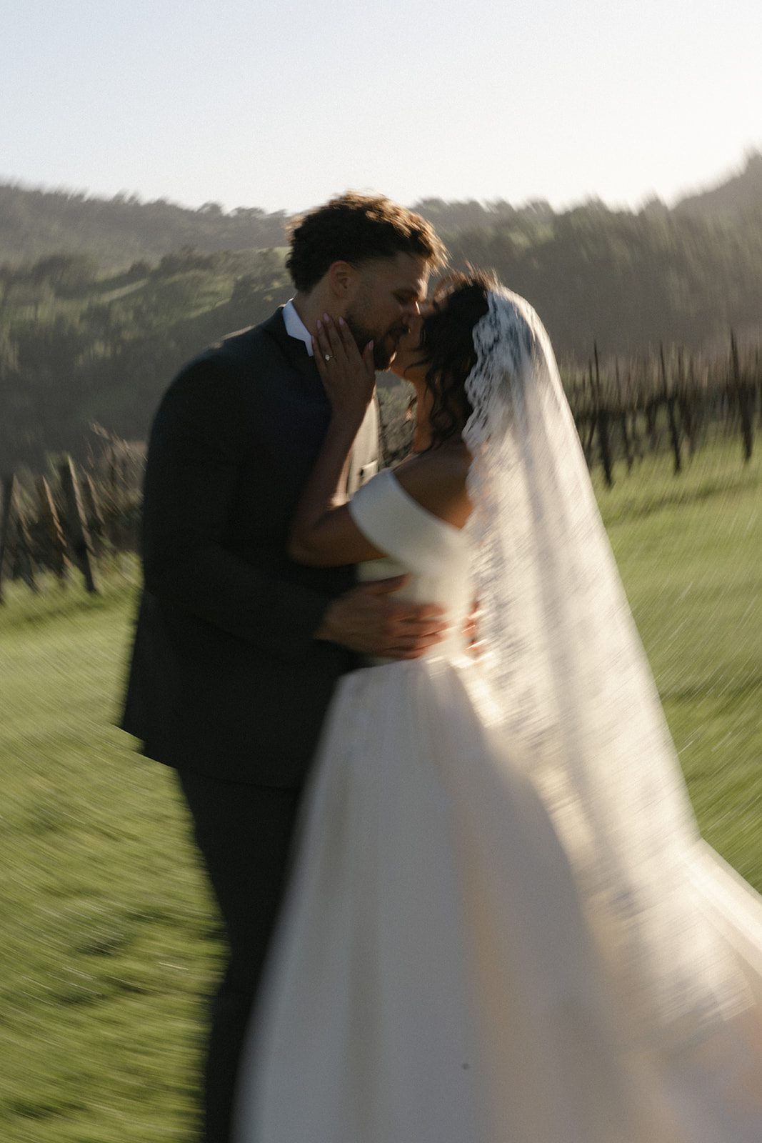 Dreamy motion-blurred wedding portrait of a bride and groom kissing in the vineyards at Sunstone Villa, featuring a flowing ivory wedding gown, delicate lace veil, and soft golden hour light over the Santa Ynez Valley.