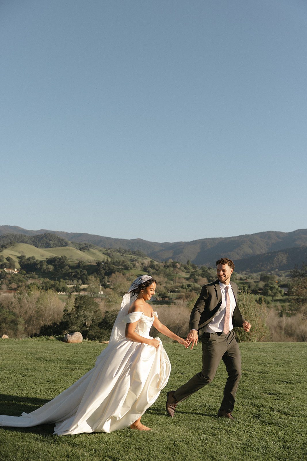 Newlyweds running barefoot across a grassy hillside at Sunstone Villa, holding hands with the rolling Santa Ynez Valley mountains stretching behind them.