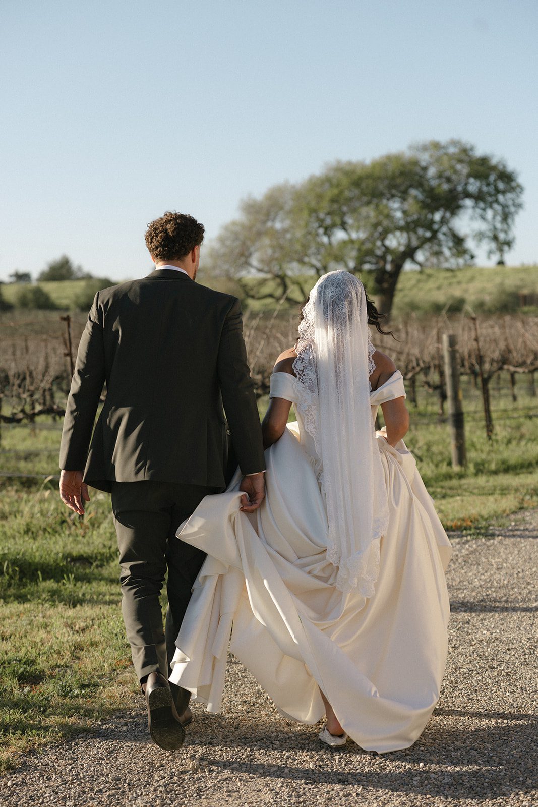Candid wedding photograph of a bride and groom walking together along a gravel path beside the vineyards at Sunstone Villa, with the bride's flowing satin gown and lace veil illuminated by warm afternoon sunlight.