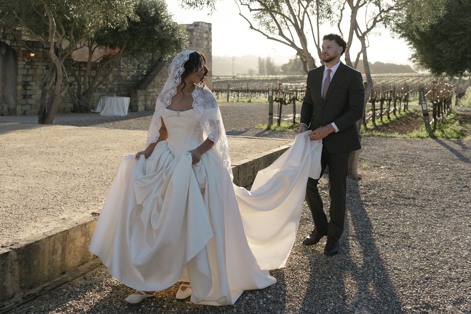 Bride in a voluminous satin wedding gown and lace veil walking along a vineyard path while her groom carries her train at a Santa Ynez wedding venue.