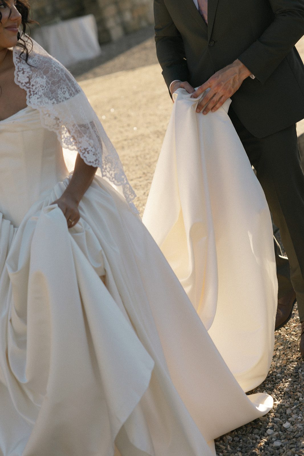 Close-up of a bride's lace veil and flowing satin wedding dress as her groom gently lifts the train during their European inspired wedding at Sunstone Villa.