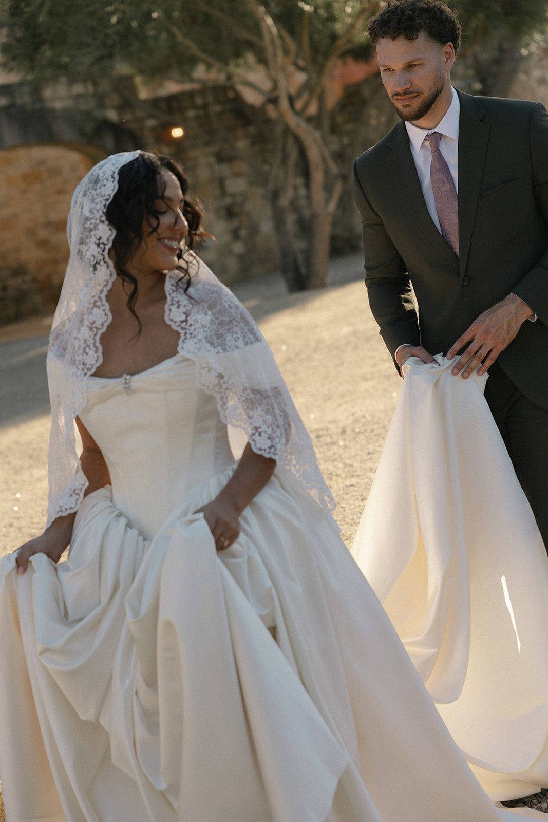 Romantic golden hour wedding portrait of a smiling bride in a lace mantilla veil and ivory satin gown, while her groom gently carries her dress train through the Sunstone Villa courtyard.
