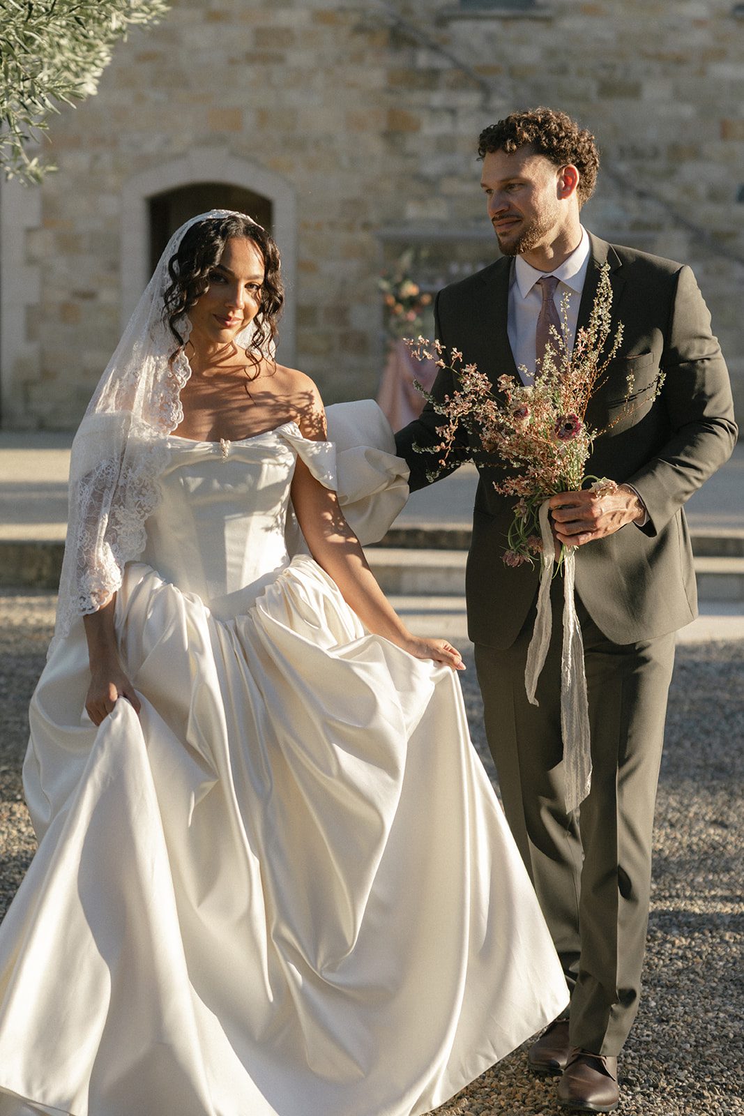 Golden hour wedding portraits outside the European inspired stone villa at Sunstone, featuring a bride in an off-the-shoulder gown and her groom holding a loose garden-style bouquet.