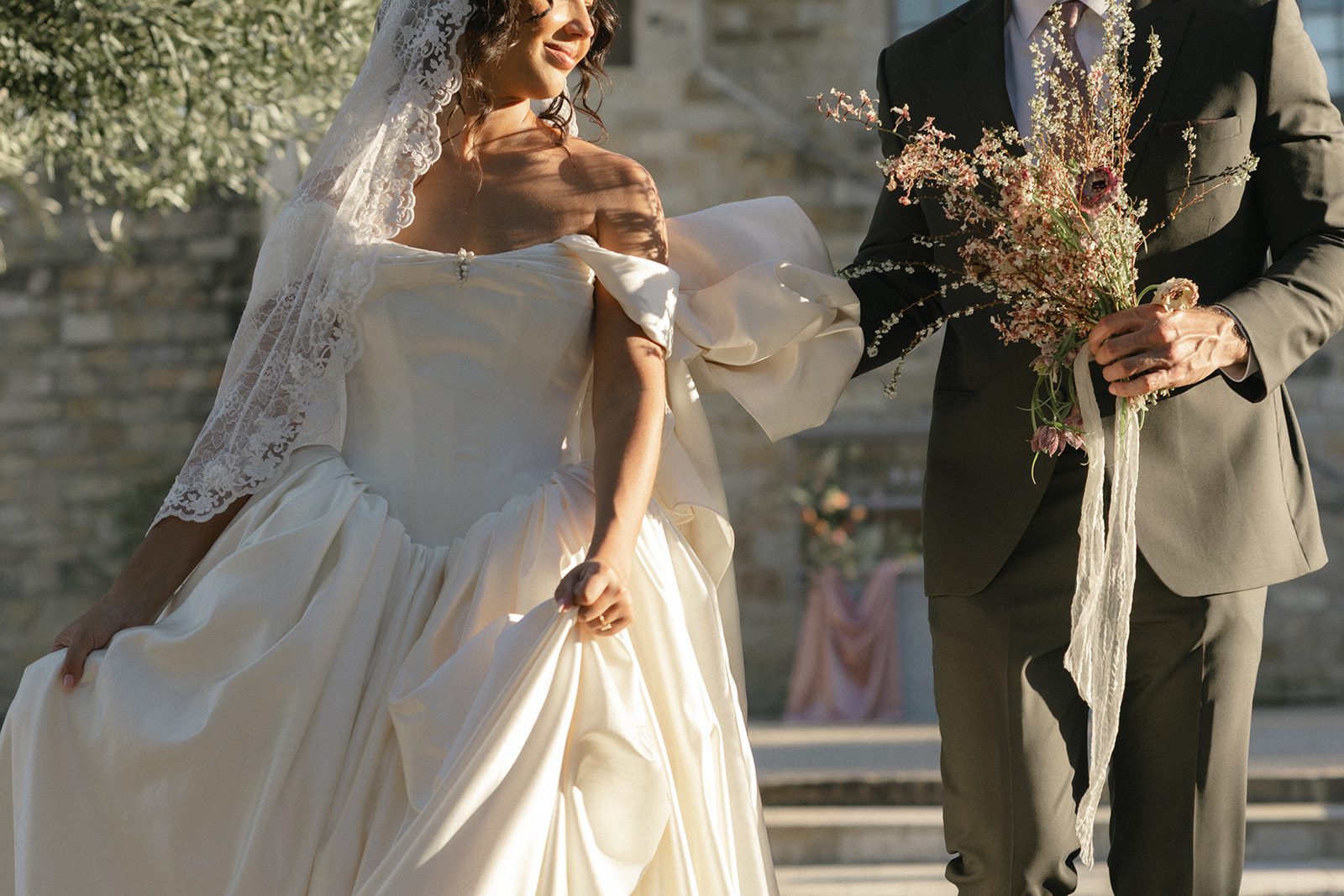 Editorial wedding detail photograph capturing a bride's elegant satin gown, lace mantilla veil, and loosely gathered skirt alongside her groom holding a delicate wildflower bouquet in soft pink and ivory tones.