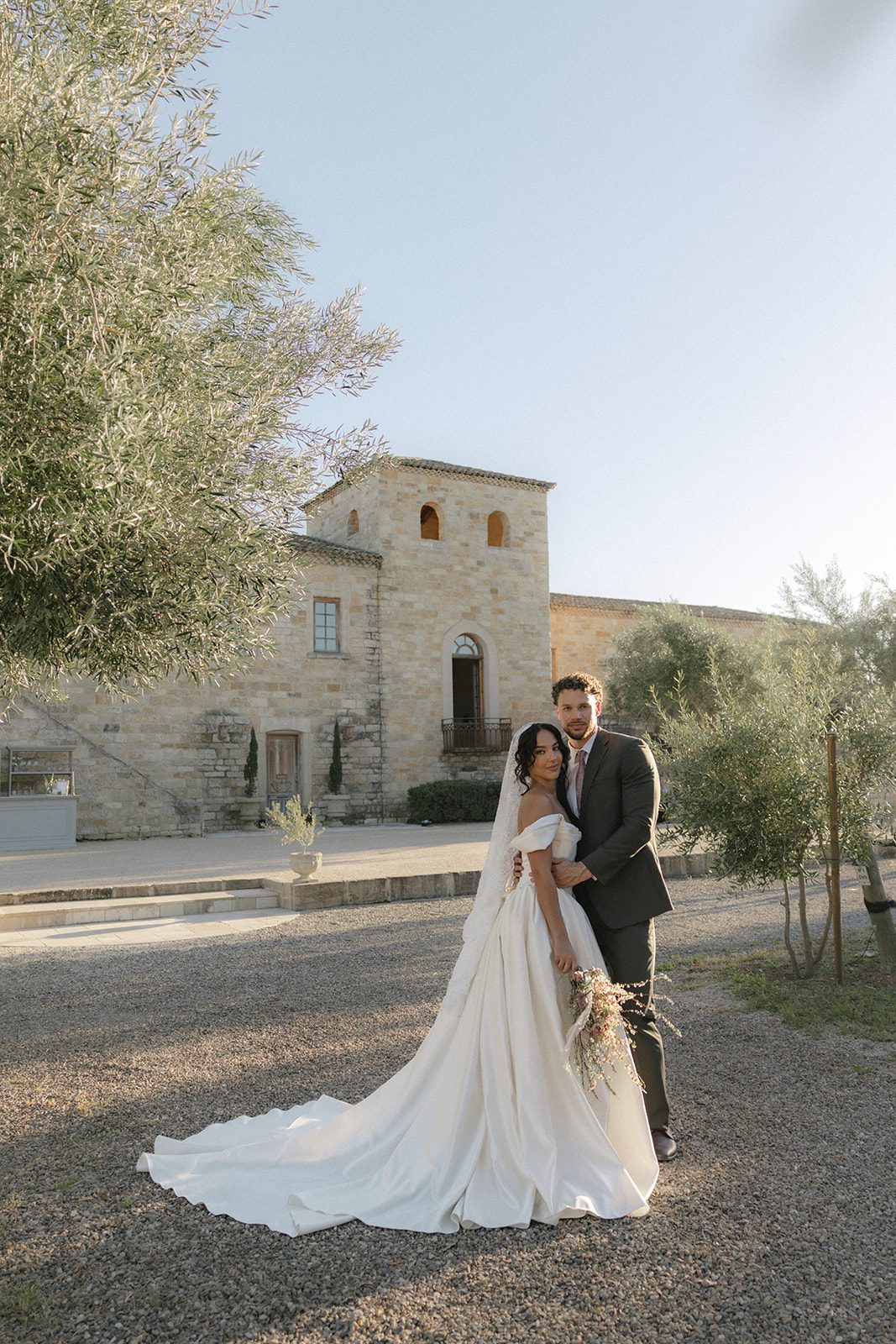 Timeless bride and groom portrait outside the French-inspired stone architecture of Sunstone Villa in Santa Ynez, featuring a romantic ivory wedding gown, lace veil, garden-style bridal bouquet, and warm golden hour light.