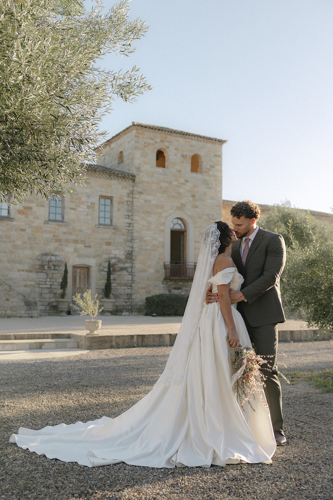 Bride and groom embracing outside the European inspired stone architecture of Sunstone Villa, with a long bridal train and delicate garden-style bouquet.