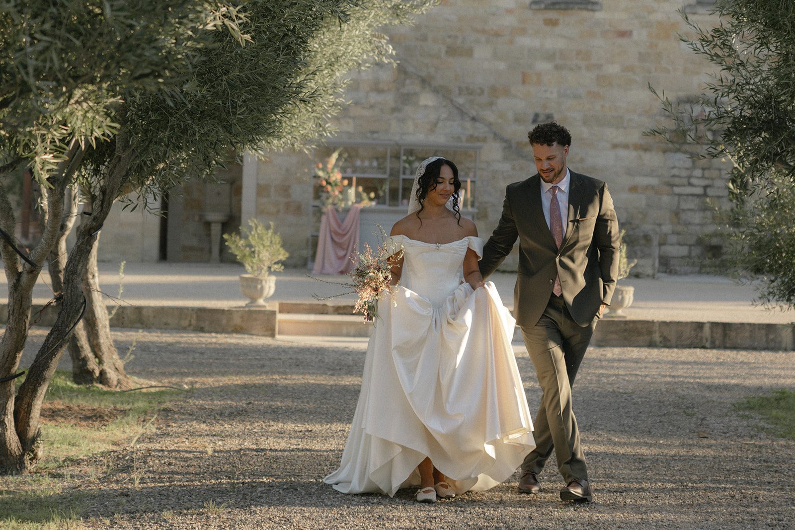 Romantic bride and groom walking together through the olive trees at Sunstone Villa in Santa Ynez, featuring an elegant off-the-shoulder satin wedding gown, garden-inspired bridal bouquet, and warm golden hour wedding photography.