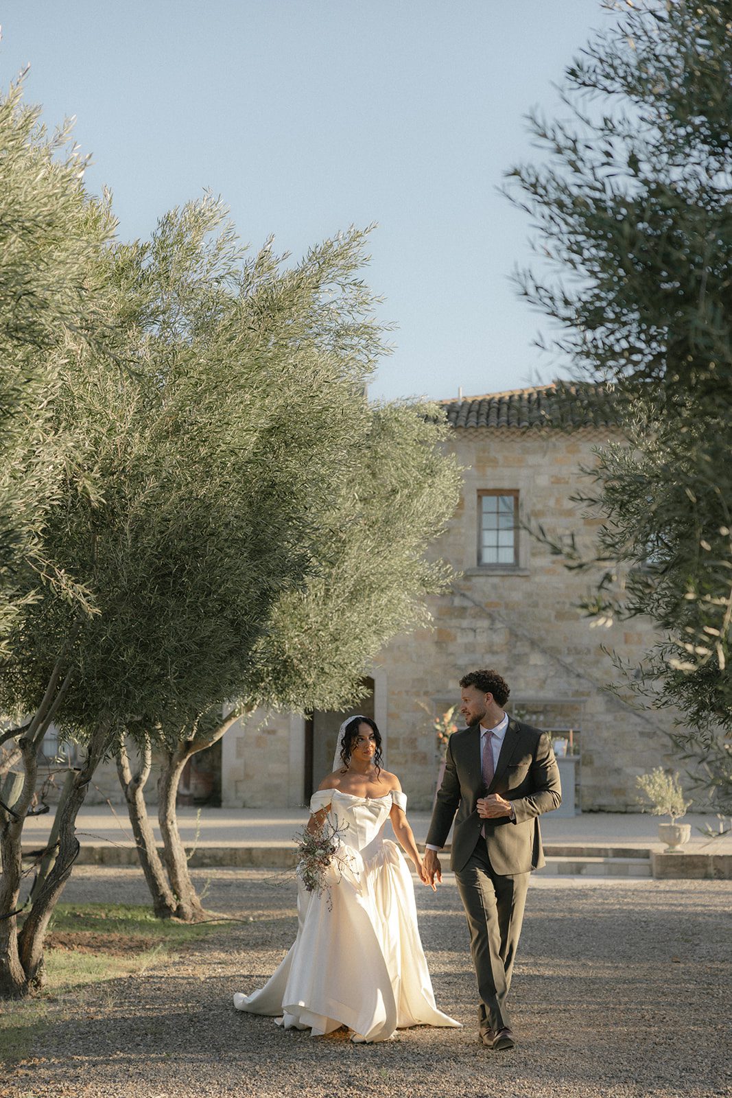 Bride and groom holding hands beneath olive trees at Sunstone Villa in Santa Ynez, surrounded by Mediterranean-inspired stone architecture and soft golden evening light.