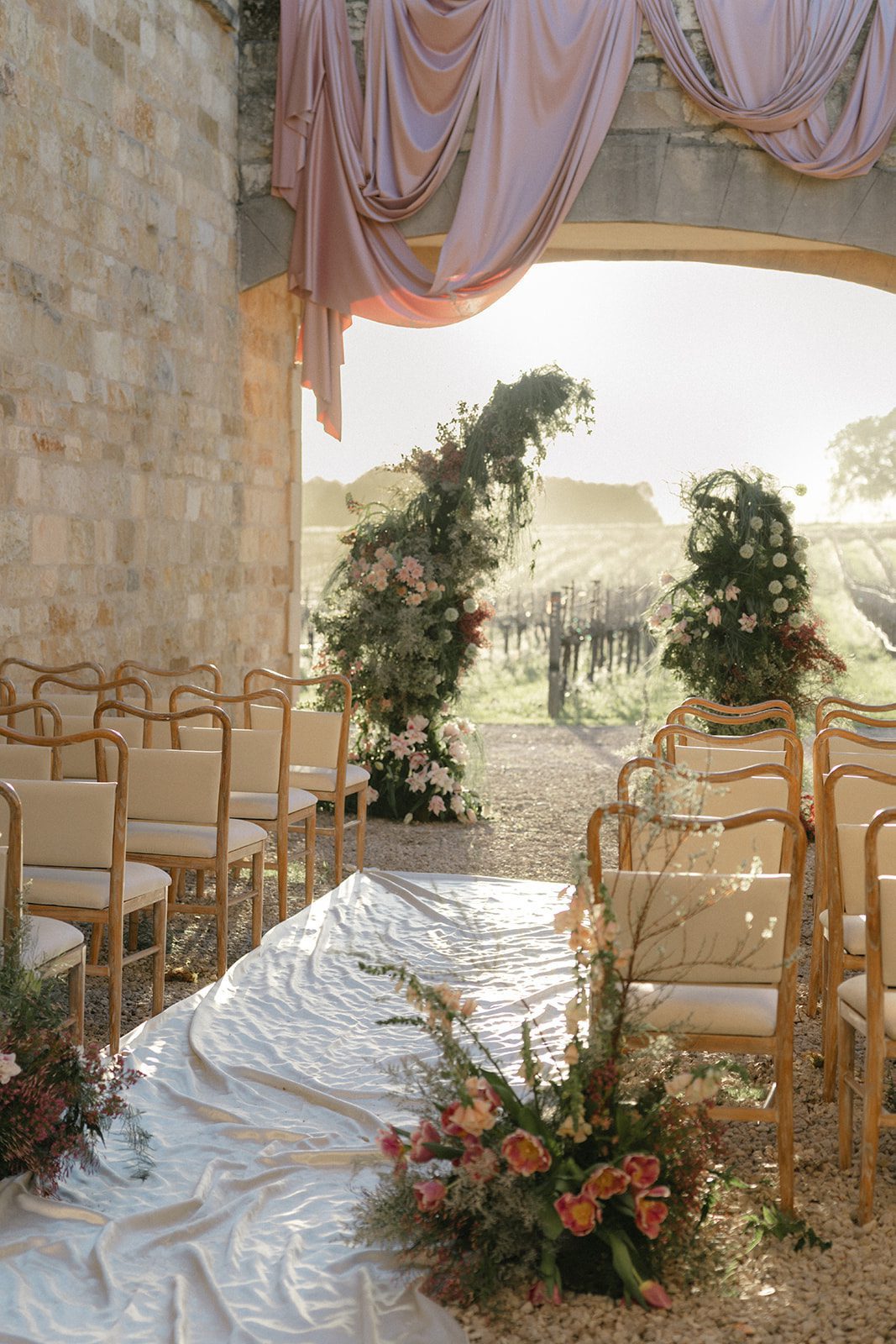 Romantic outdoor wedding ceremony setup at Sunstone Villa in Santa Ynez, featuring blush pink fabric draped over a limestone archway, lush garden-style floral installations, a flowing ivory aisle runner, and vineyard views illuminated by golden sunlight.