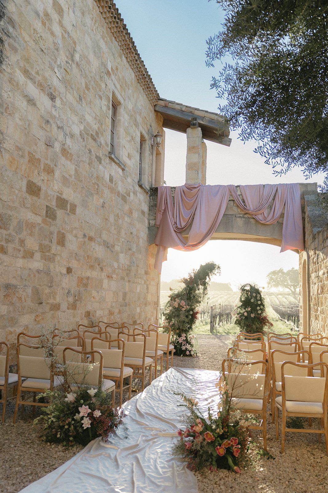 Intimate outdoor wedding ceremony setup at Sunstone Villa, featuring a flowing ivory aisle runner, blush pink draping across a stone archway, lush garden-style floral installations, and wooden chairs overlooking the vineyards.