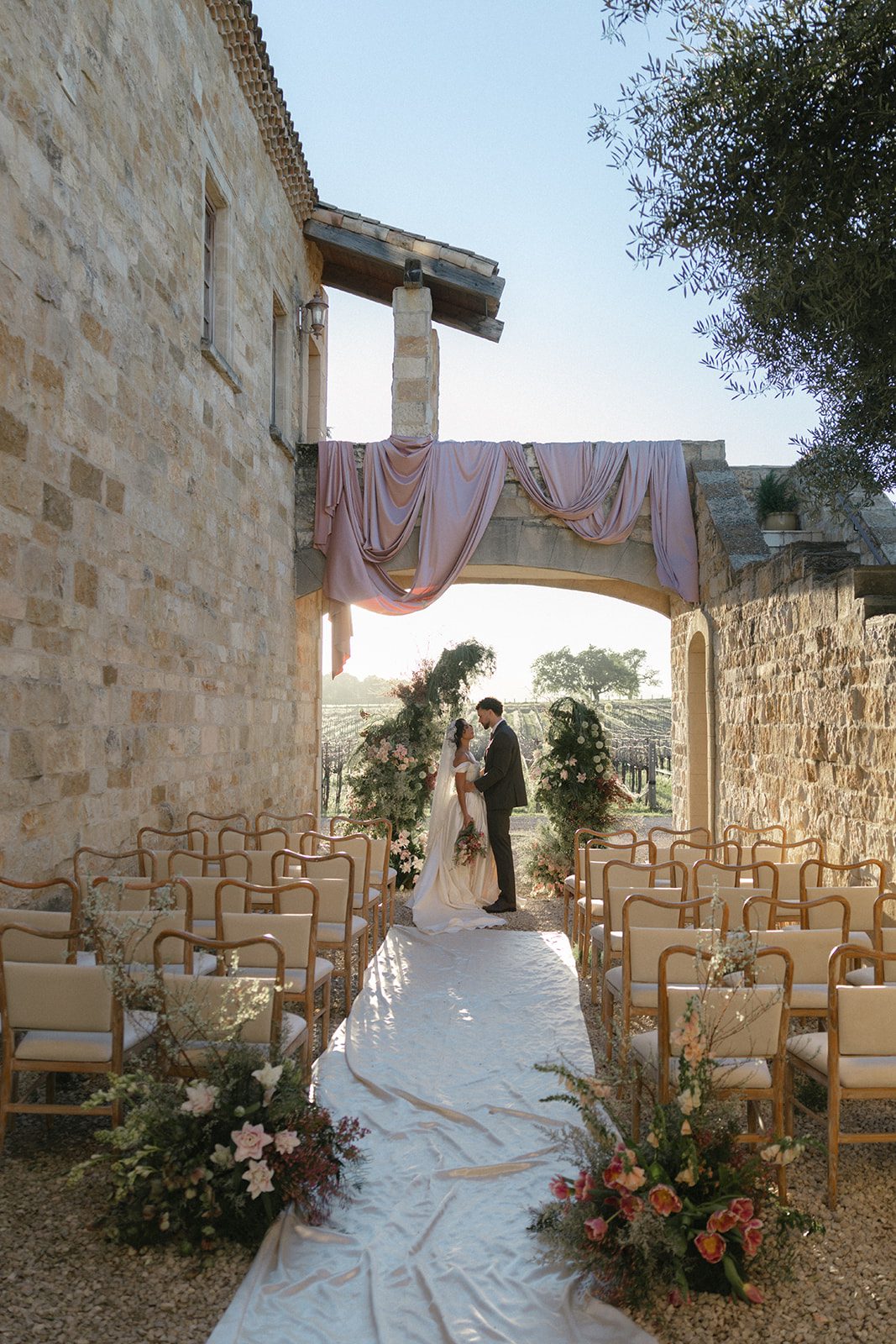 Romantic vineyard wedding ceremony at Sunstone Villa featuring a draped stone archway, lush garden florals, wooden chairs, and a fabric-lined aisle overlooking the vines.