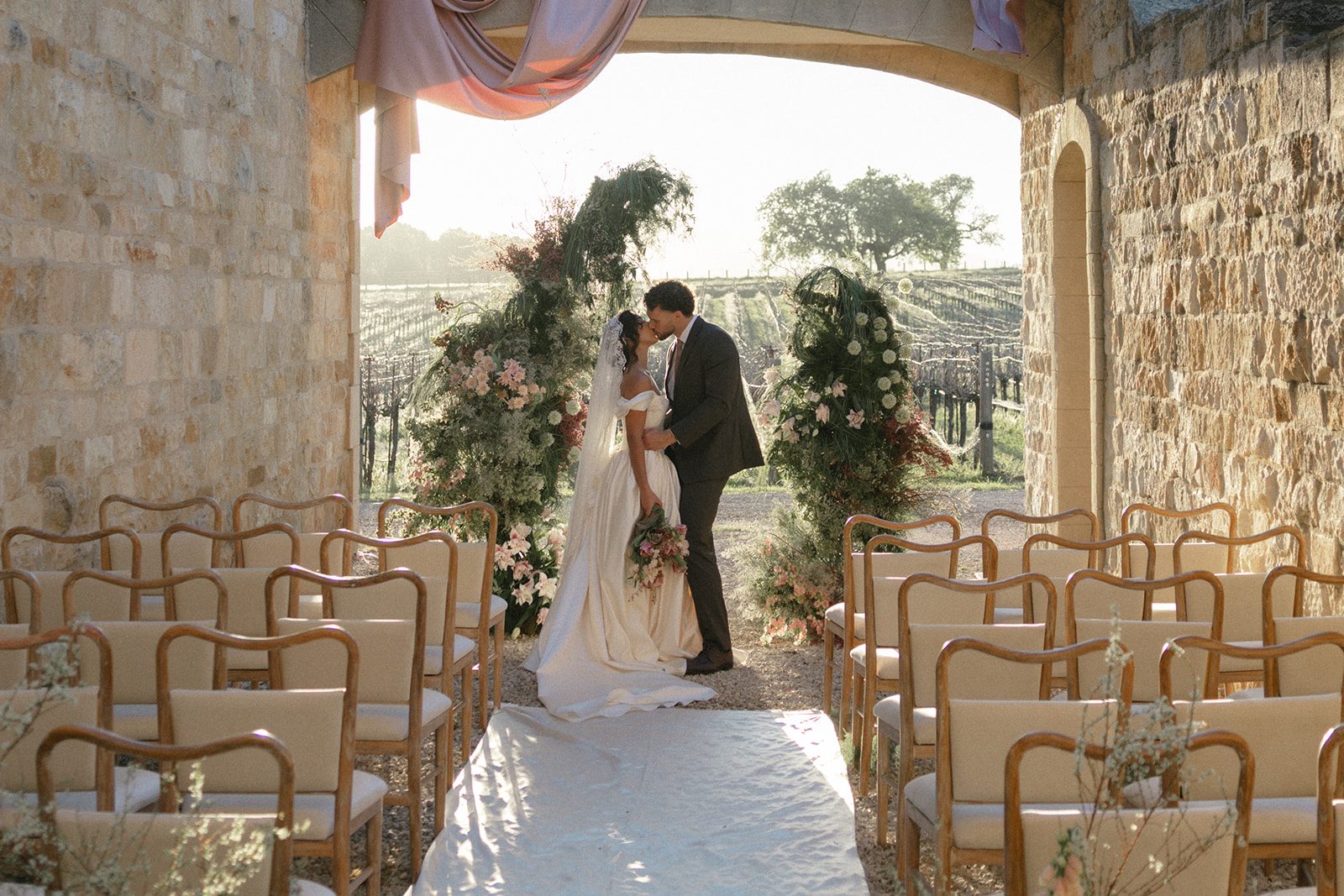 Romantic vineyard wedding ceremony at Sunstone Villa, featuring a bride and groom kissing beneath a stone archway adorned with blush pink draping and lush floral installations, surrounded by wooden ceremony chairs and golden California sunlight.