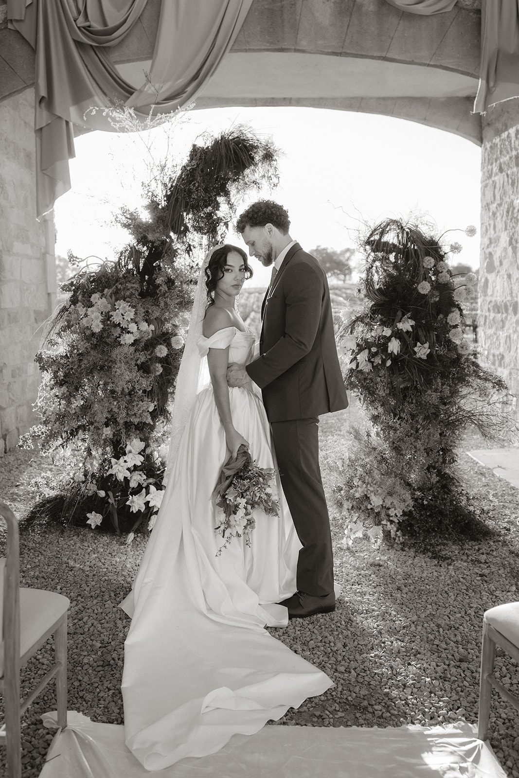 Black and white wedding portrait beneath a draped stone archway at Sunstone Villa, with the bride and groom surrounded by abundant floral installations and vineyard views.