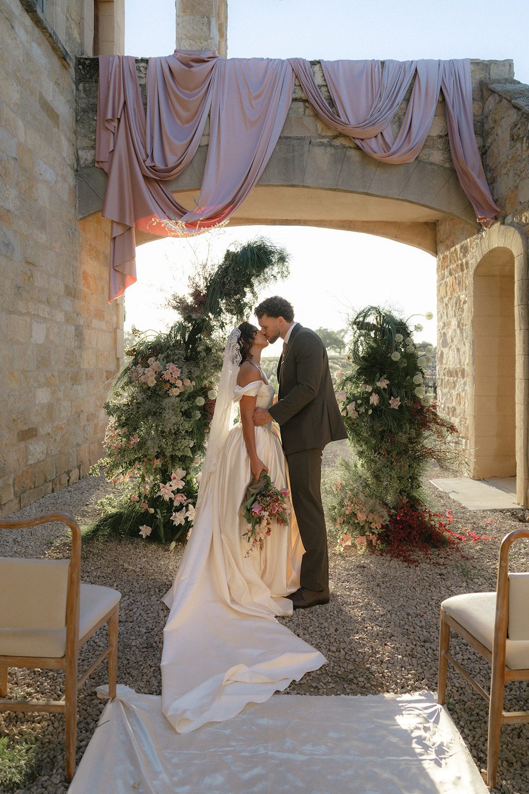 Intimate wedding portrait of a bride and groom beneath a romantic limestone archway at Sunstone Villa, featuring cascading garden-style floral installations, blush pink drapery, an ivory satin wedding gown, and warm California sunlight.