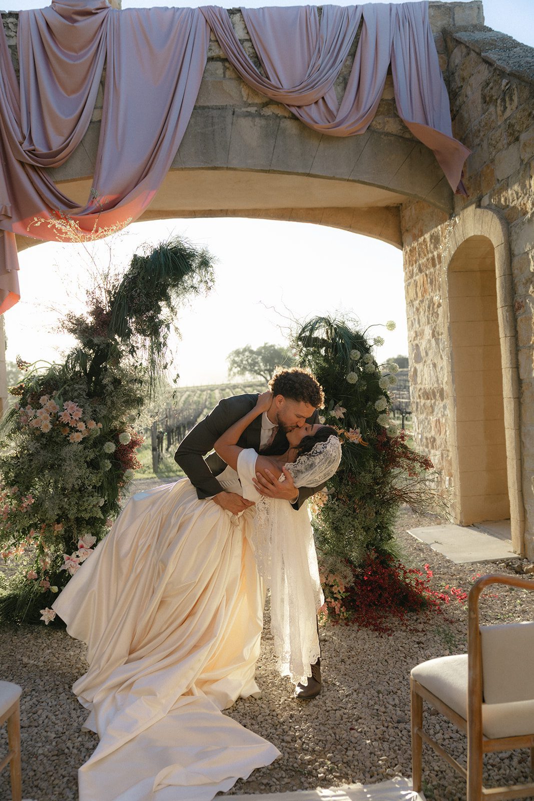 Groom dipping and kissing his bride beneath a blush-draped stone arch surrounded by lush garden florals during their Sunstone Villa Wedding ceremony, photographed in golden hour light.