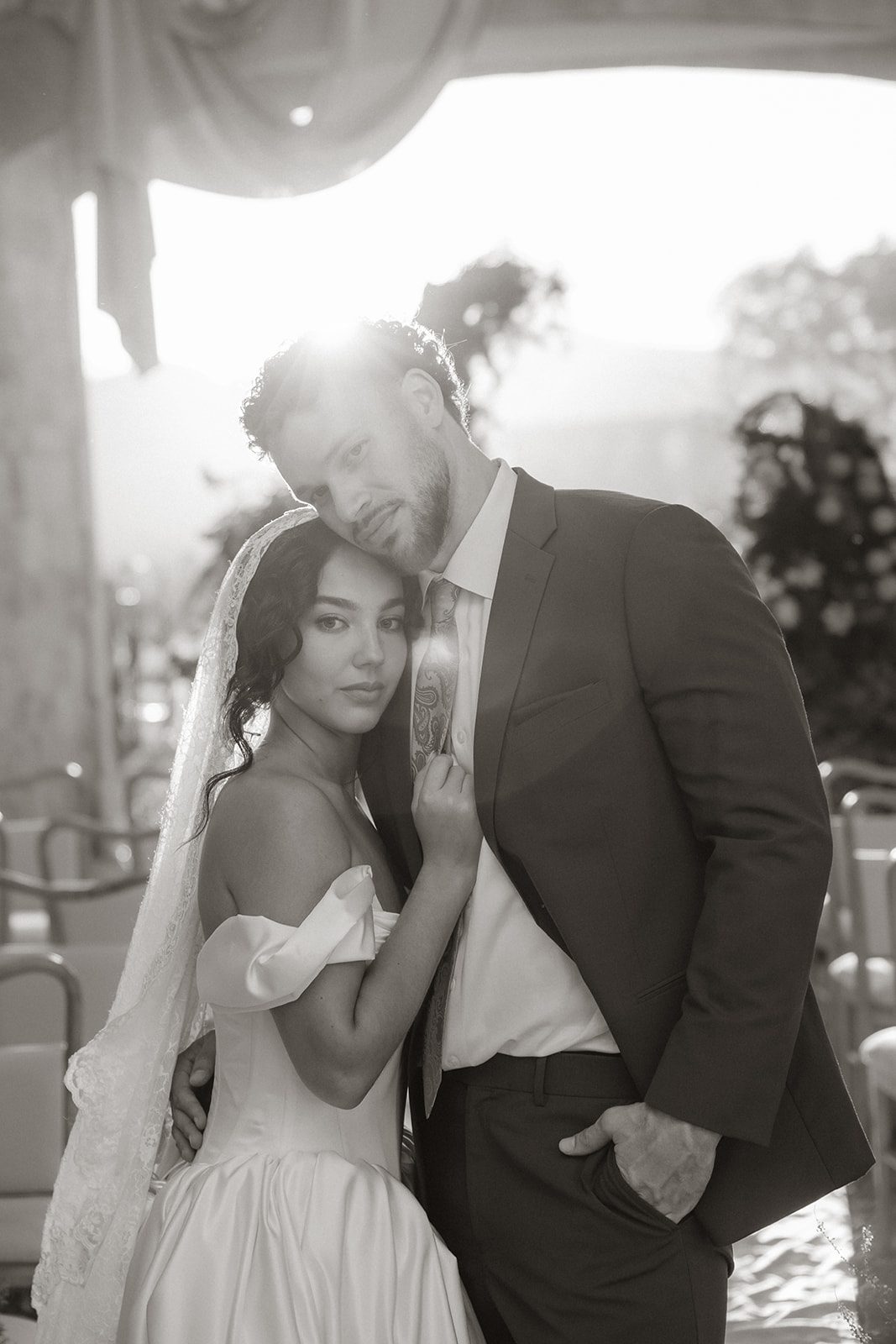 Black and white wedding portrait captured by a California wedding photographer, featuring a bride in a lace veil leaning into her groom beneath the stone archway of Sunstone Villa.
