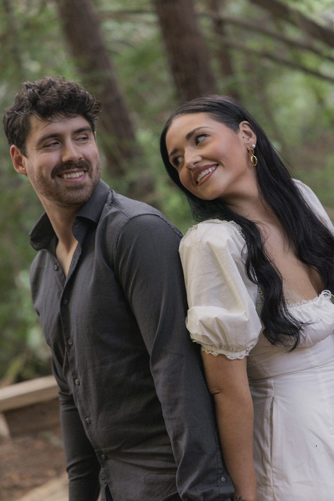 Couple leaning back-to-back and smiling during a forest session with a Bay Area wedding photographer.
