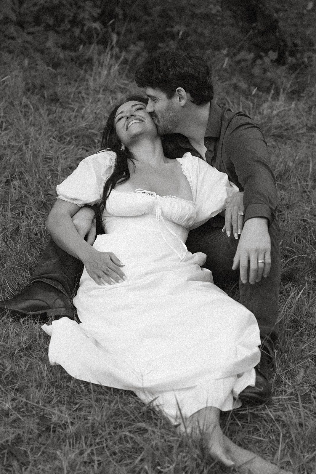 Black-and-white portrait of an engaged couple sitting in the grass as one kisses the other’s cheek, a candid moment from Northern California engagement photos.