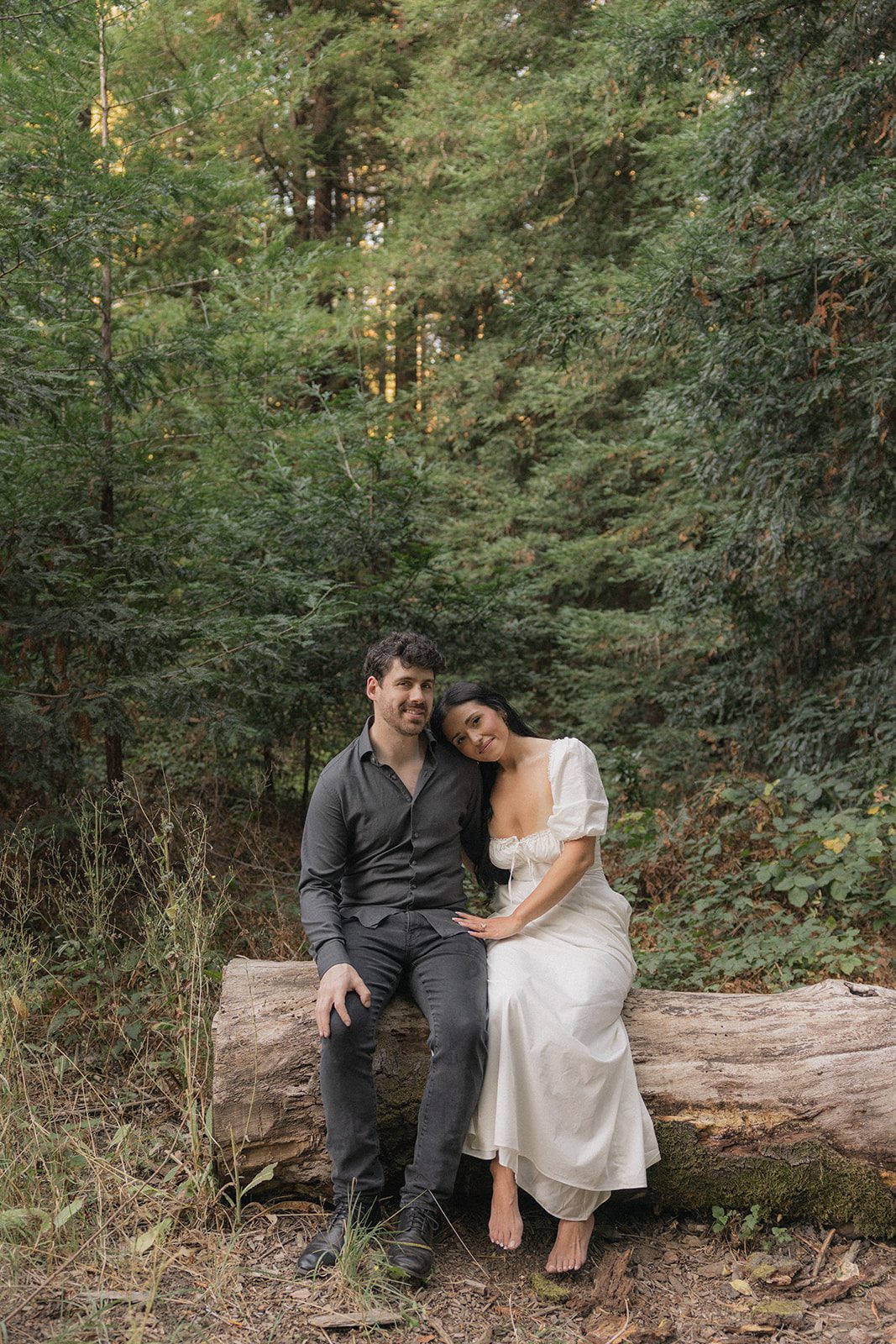 Couple sitting together on a fallen redwood log surrounded by dense greenery for Reinhardt Redwood Regional Park Engagement Photos.