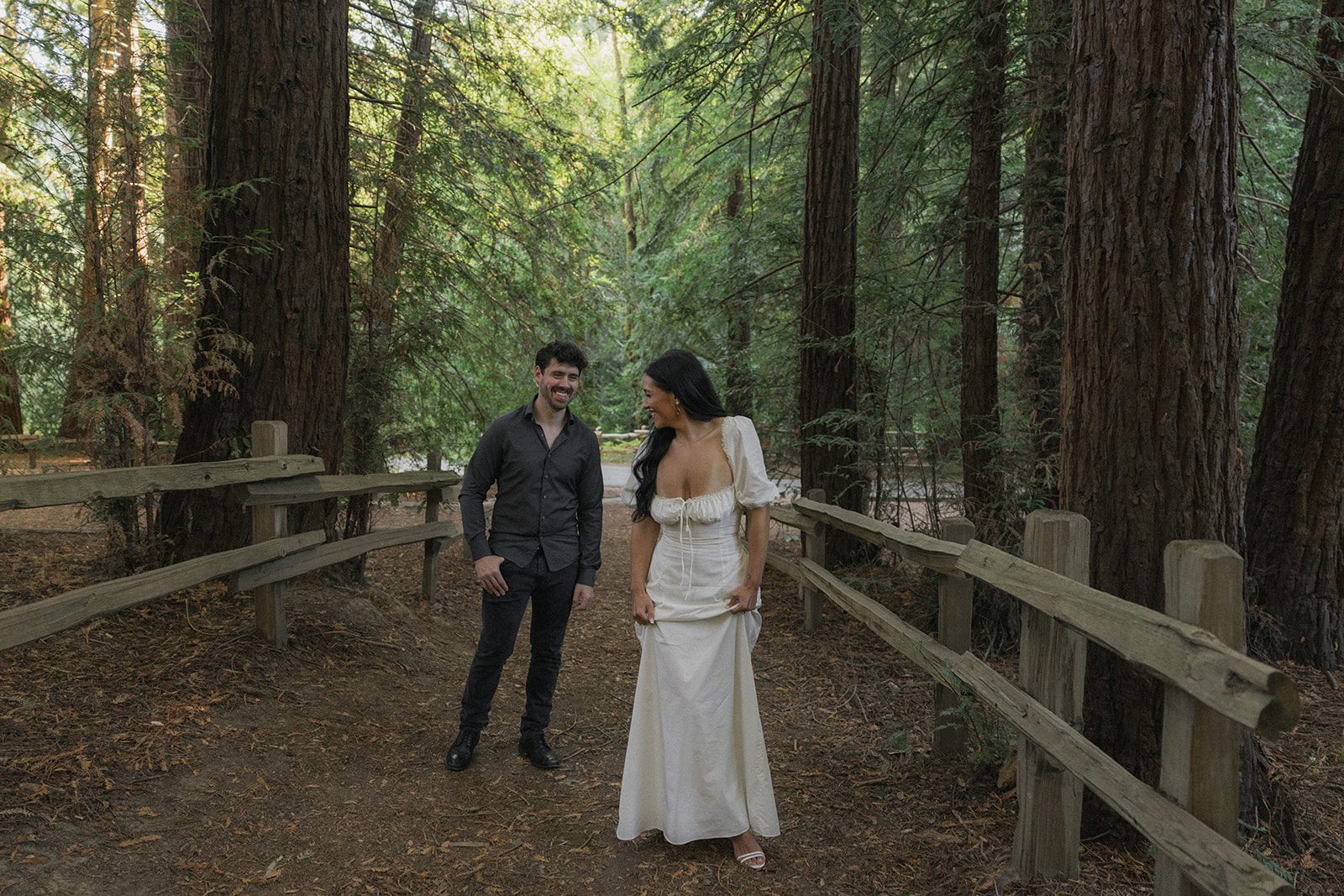 Couple laughing together on a fenced trail surrounded by towering redwoods, natural forest engagement photos by a Bay Area wedding photographer.