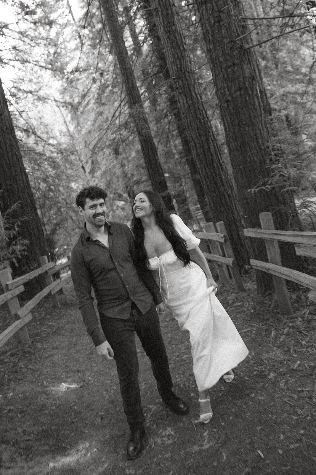Black-and-white portrait of an engaged couple laughing together while walking beneath towering redwoods, captured during Reinhardt Redwood Regional Park Engagement Photos.