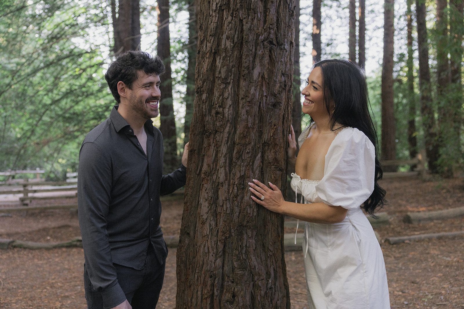 Couple smiling at each other from opposite sides of a towering redwood tree, highlighting one of the scenic engagement photo locations in the Bay Area for Northern California engagement photos.