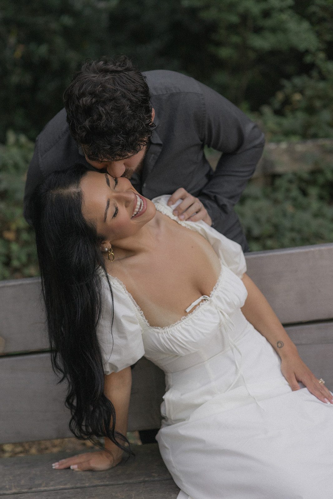 Engaged couple sharing a playful moment on a wooden bench surrounded by greenery, photographed during Reinhardt Redwood Regional Park Engagement Photos.