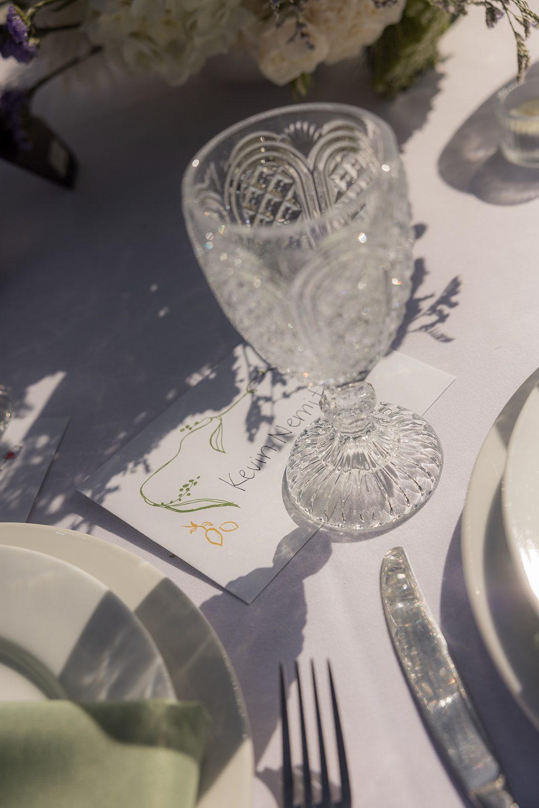 Vintage cut-glass goblet resting on a personalized place card beside white dinnerware and a sage green napkin at a vineyard wedding venue.
