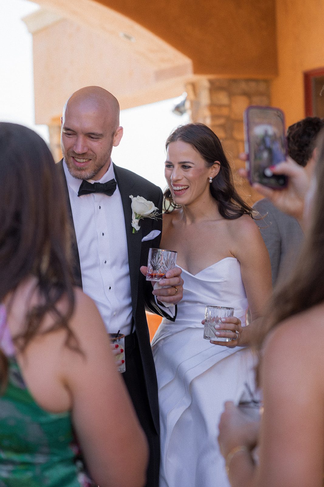 Bride in a strapless white gown and groom in a black tuxedo mingle with guests while holding drinks at their Villa San Juliette wedding reception.