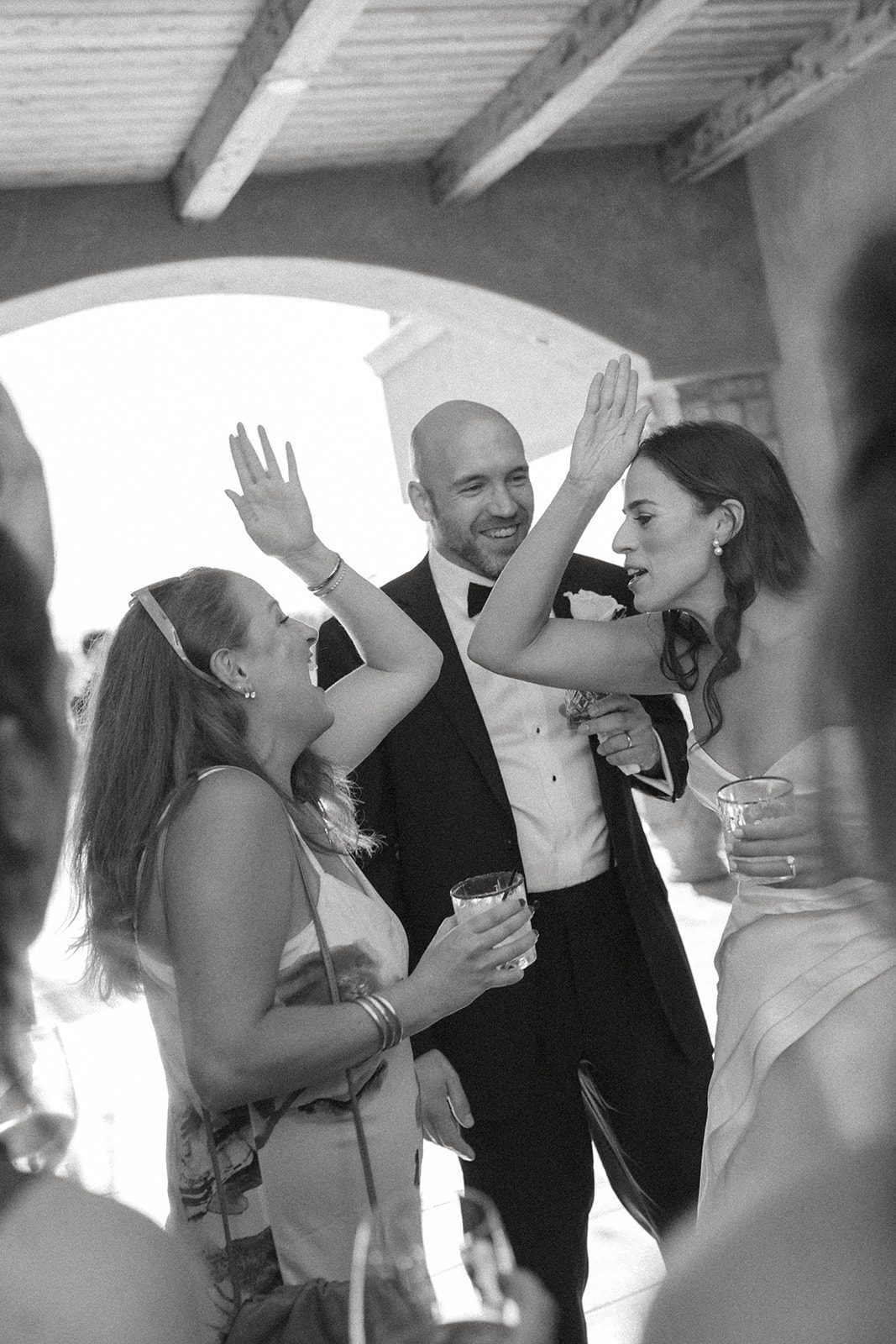Black-and-white photograph of a bride high-fiving a guest while the groom smiles during their Villa San Juliette wedding cocktail hour.
