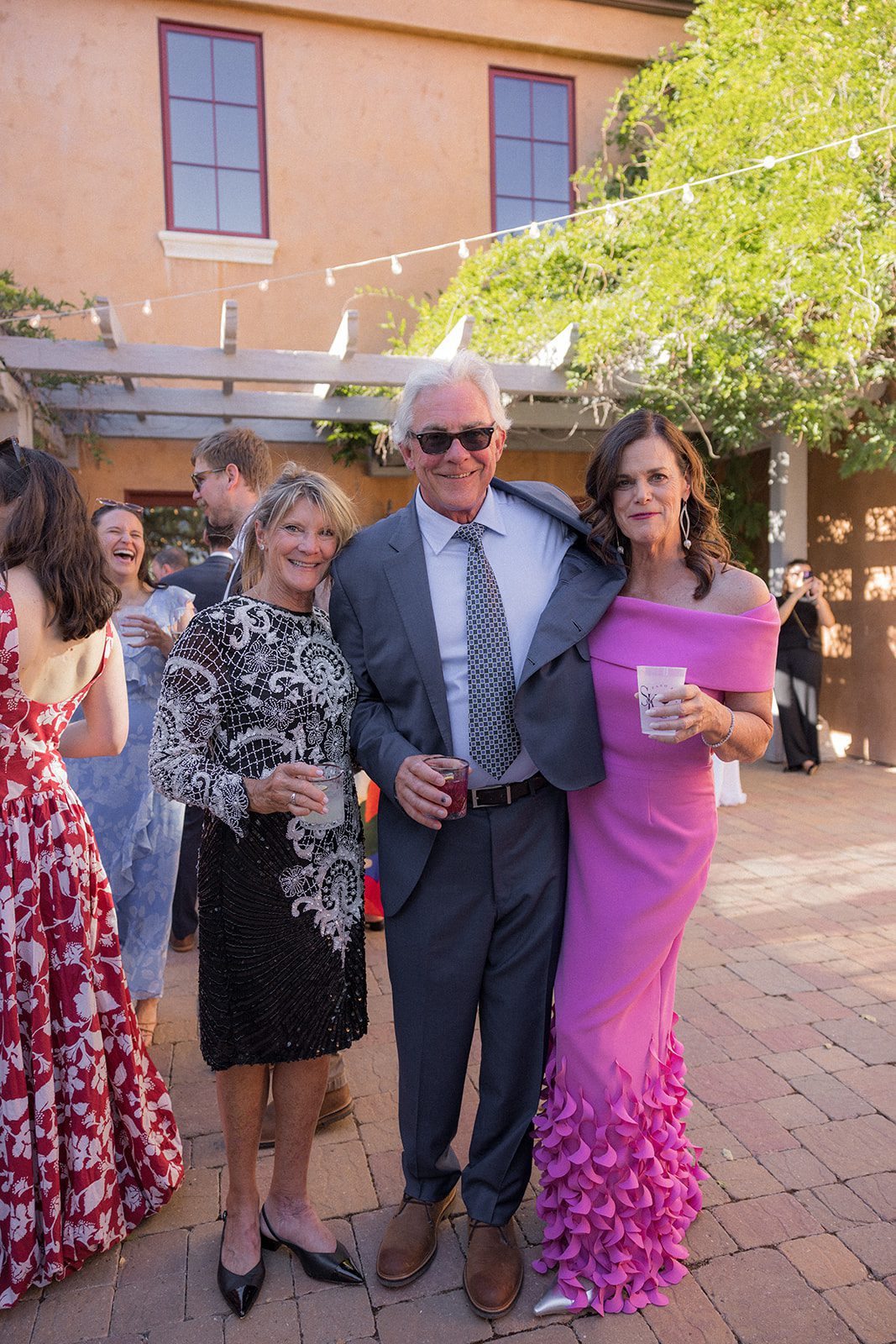 Three wedding guests pose with drinks in a sunlit courtyard, wearing a black embroidered dress, gray suit, and bright pink gown at Villa San Juliette.