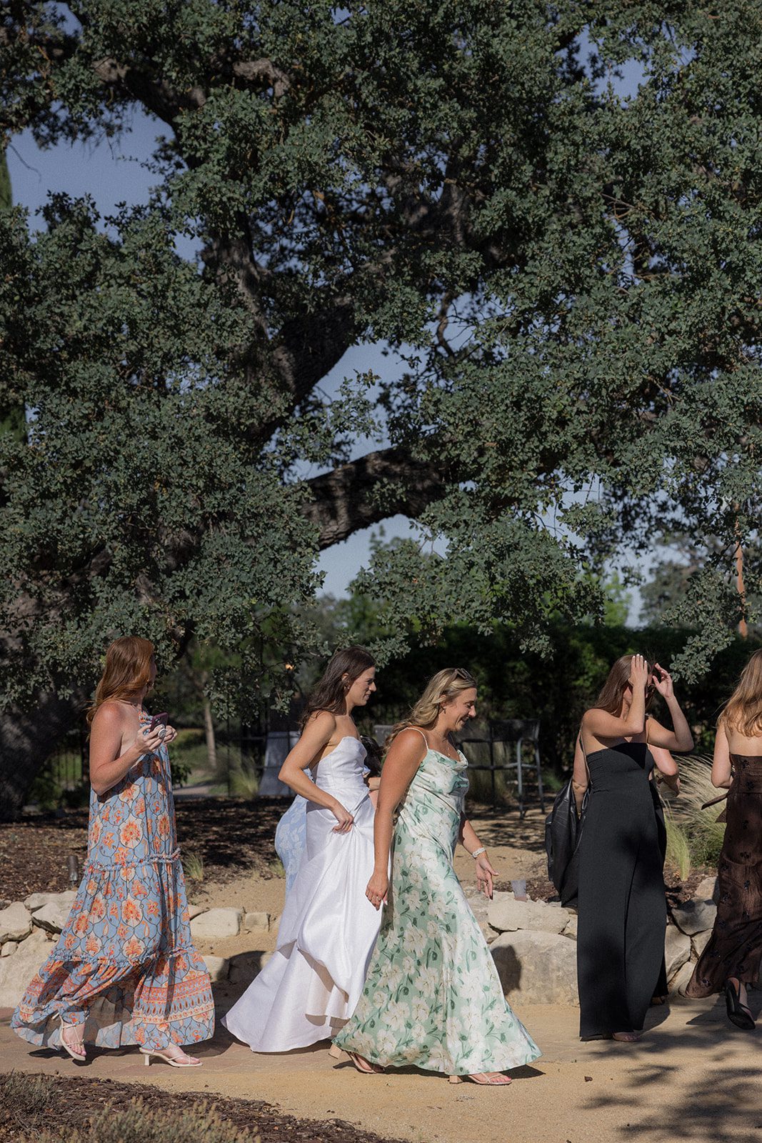 Bride walking alongside guests in colorful summer dresses beneath a large oak tree during a san luis obispo wedding.