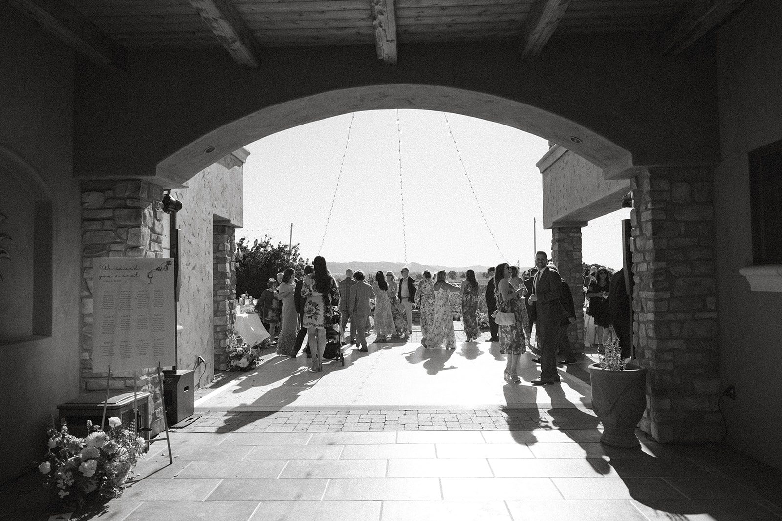 Black-and-white view through a stone archway of guests gathering beneath string lights at a vineyard wedding venue near San Luis Obispo.
