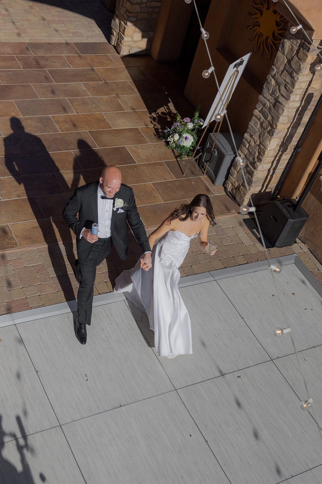 Overhead photograph of newlyweds holding hands as they enter the outdoor dance floor at their vineyard wedding venue beneath string lights.